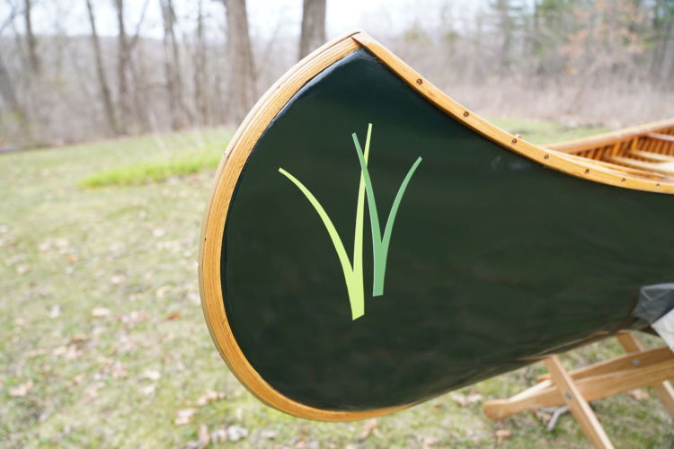 The bow of a dark green wooden canoe with two pairs of grassy green leaves from the logo of the Wisconsin Wetlands Association.