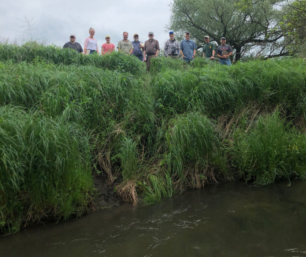 Photo taken from the stream level looking towards the very steep and tall streambanks with a group of people at the very top for scale.