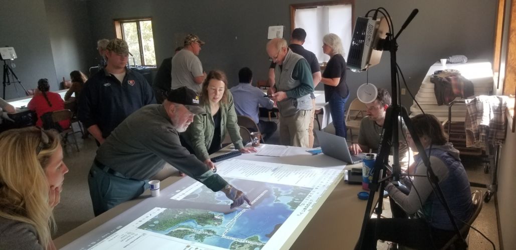 A group of workshop attendees surrounds a map projected onto a table, with two people pointing to a feature on the map.