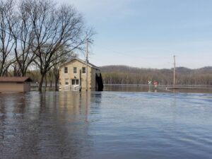High water makes up most of the foreground, rising up to the front door of a historic building in the background and partially up a Stop sign and Railroad crossing sign in the distance. Presumably, the photographer is standing on a road that is now covered in water.