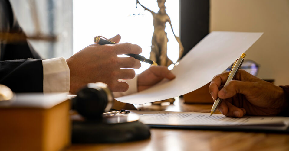 A photo of two sets of hands looking through papers and gesturing with pens, with a statue of the 'Scales of Justice' in the background.